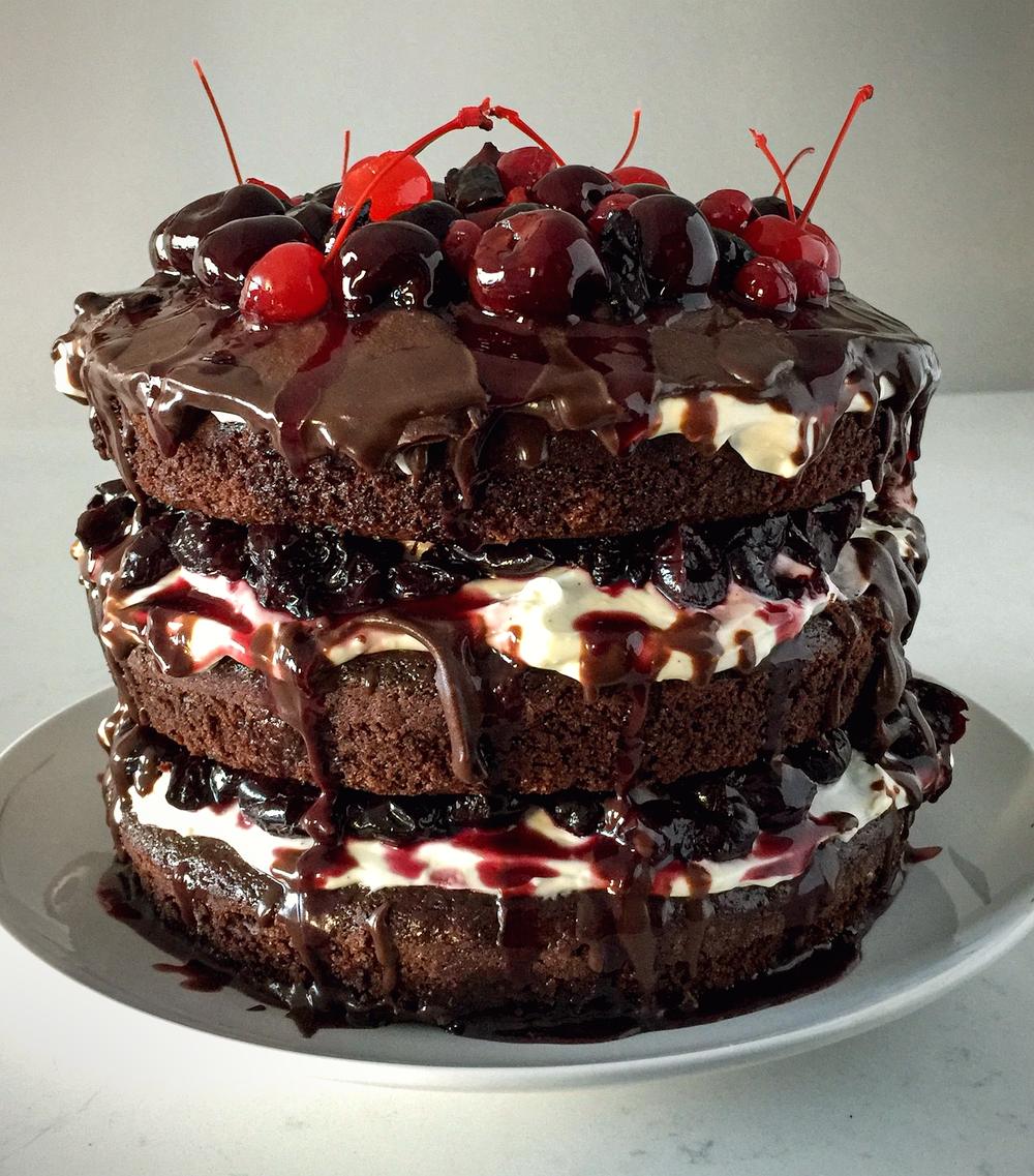 A three-layer black forest cake, unfrosted at the sides so the sponge, cream and cherry layers show, with dark ganache and cherries piled on top.
